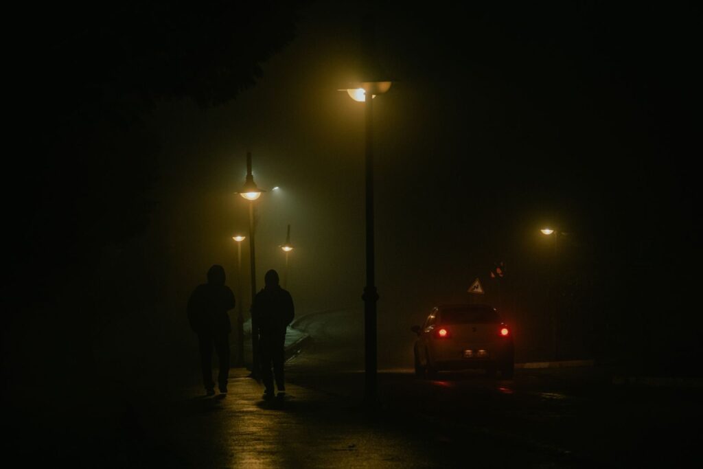 Two people walk on a foggy street at night, illuminated by streetlights.